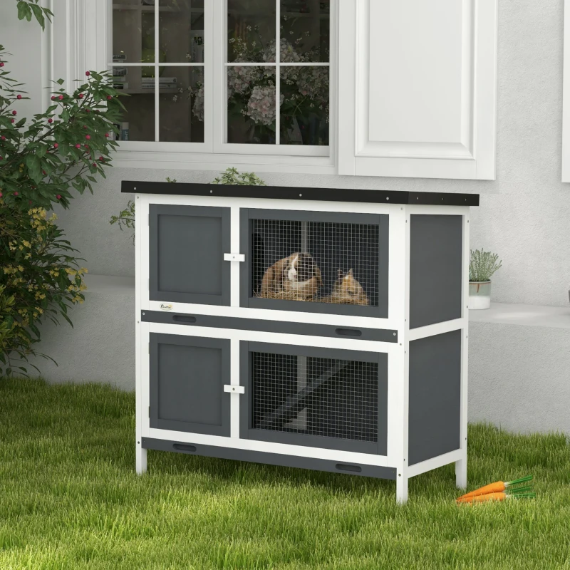 Two-level grey and white rabbit hutch outdoors with two rabbits inside the top section.
