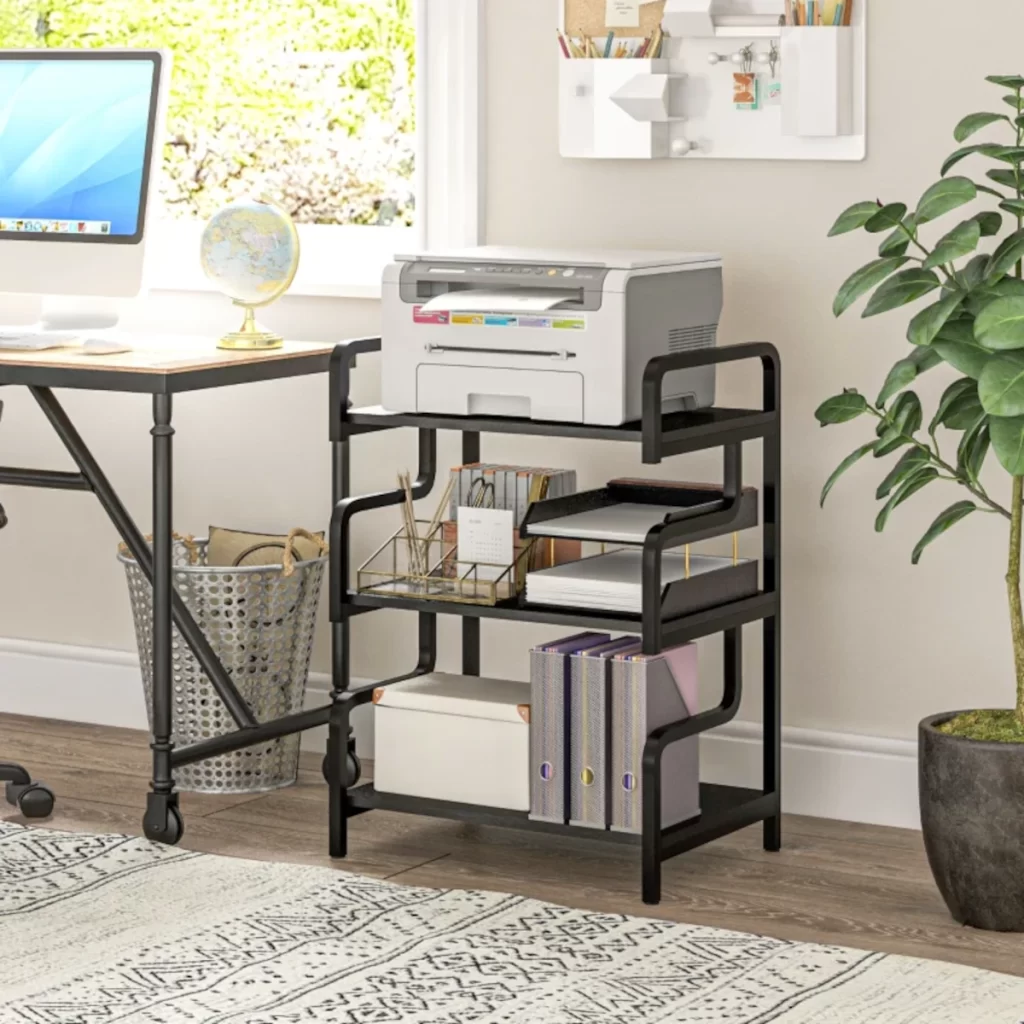 Three-tier black storage shelf holding a printer, books, files, and office accessories in a home workspace.