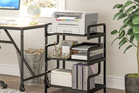 Three-tier black storage shelf holding a printer, books, files, and office accessories in a home workspace.